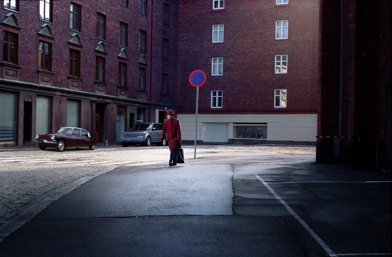 A woman in a red coat and hat carries a black bag across a cobblestone courtyard past a vintage red car and a no-parking sign.