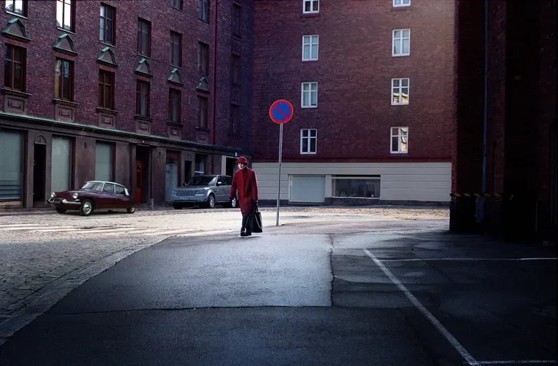 A woman in a red coat and hat carries a black bag across a cobblestone courtyard past a vintage red car and a no-parking sign.