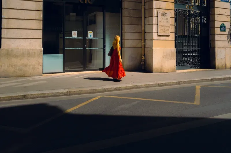 A woman in a long red dress walks alone past the stone facade of a bank, lit by low sun above a road lying in deep shadow.