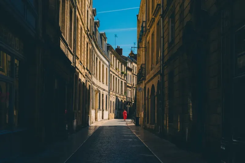 A woman in a red polka-dot dress walks alone down a quiet, sunlit stone alley lined with historic facades.