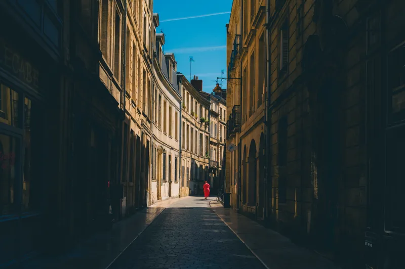 A woman in a red polka-dot dress walks alone down a quiet, sunlit stone alley lined with historic facades.