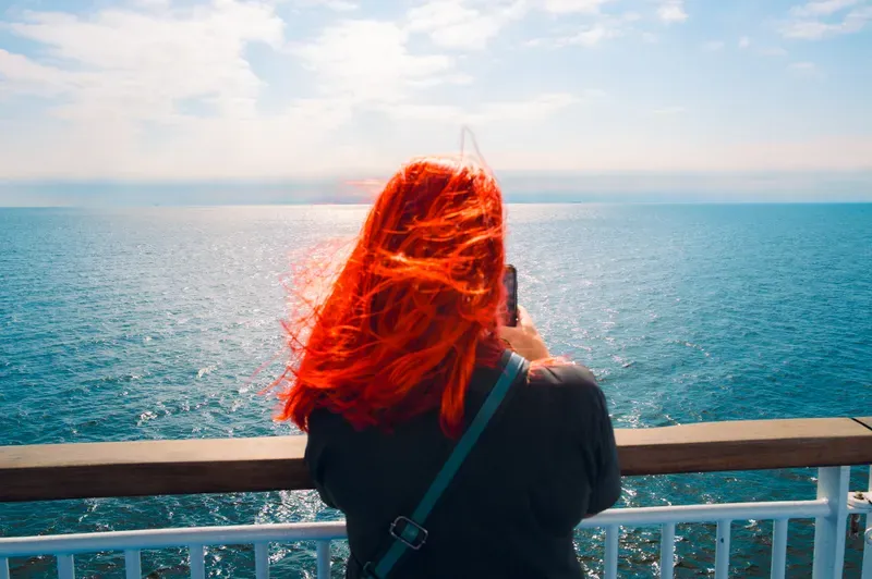 Seen from behind, a woman with vivid red hair stands at a ship's railing, raising her phone to photograph the open sea.