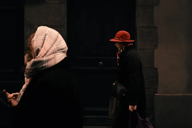 A woman in a red hat and quilted coat passes another in a plaid headscarf in a dark alley, warm light catching their faces.