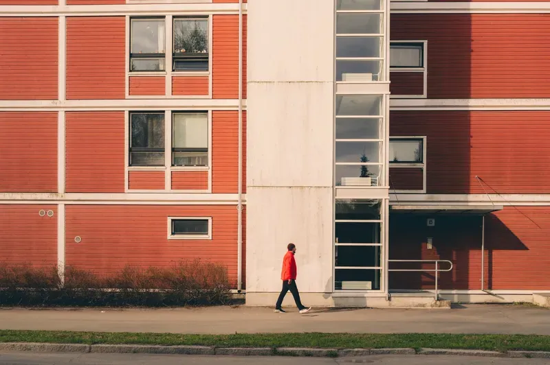 A man in a red jacket and beanie walks along a red timber-clad apartment building beside a white concrete stairwell column.