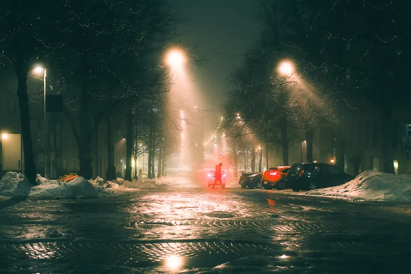 A person walks beside a car's glowing red taillights on a wet street lined with Christmas-lit trees and parked cars.