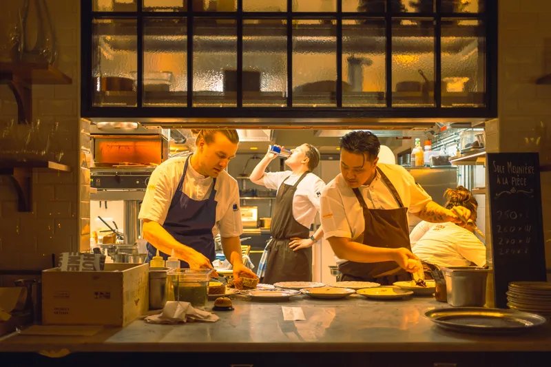 Chefs plate dishes side by side in a busy open restaurant kitchen during evening service, a chalkboard menu listed beside them.