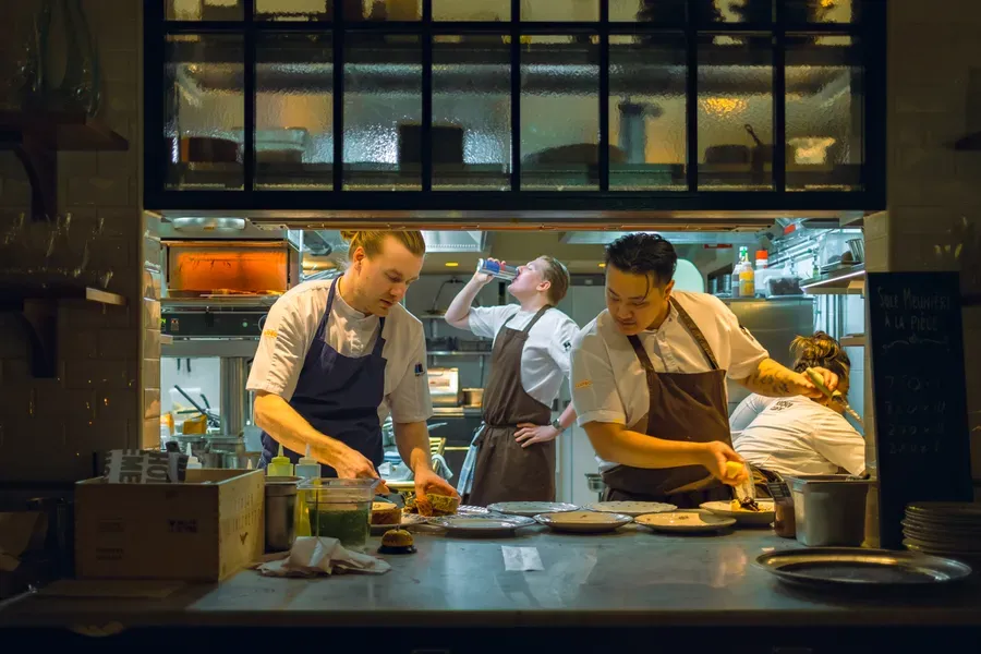 Chefs plate dishes side by side in a busy open restaurant kitchen during evening service, a chalkboard menu listed beside them.