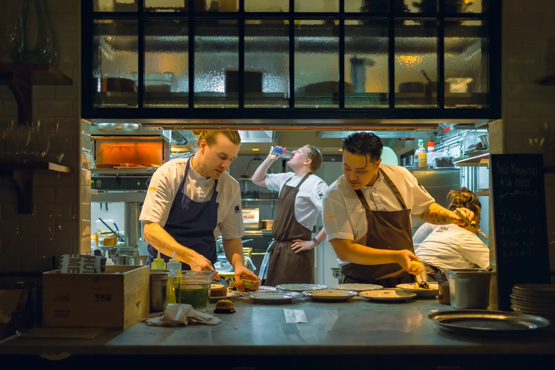 Chefs plate dishes side by side in a busy open restaurant kitchen during evening service, a chalkboard menu listed beside them.