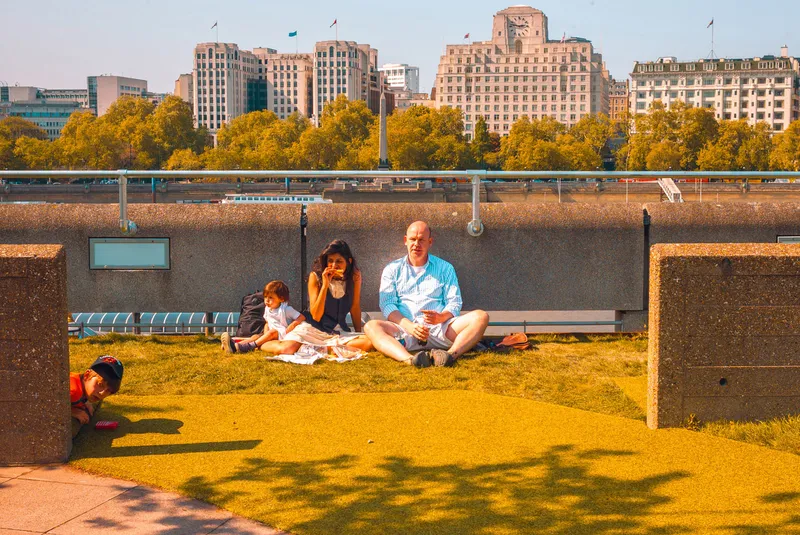 A family sits on a riverside lawn having a picnic, a boy peeking from behind a concrete wall in the foreground, a city skyline across the water.