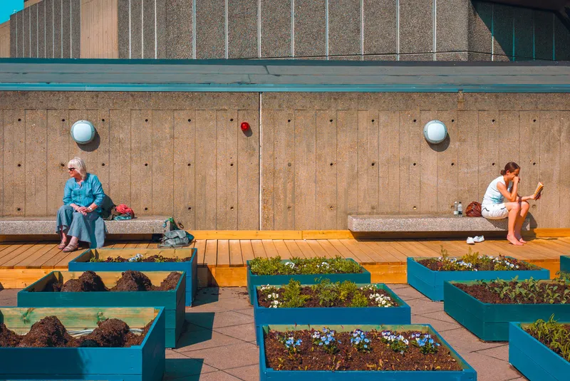Two women sit apart on concrete benches beside raised flower beds on a rooftop garden terrace.