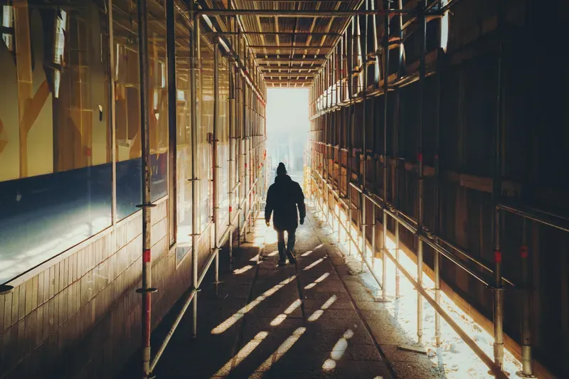 A man walks through a scaffolding-covered walkway, bright daylight streaming in and casting diagonal stripes of light across the ground.