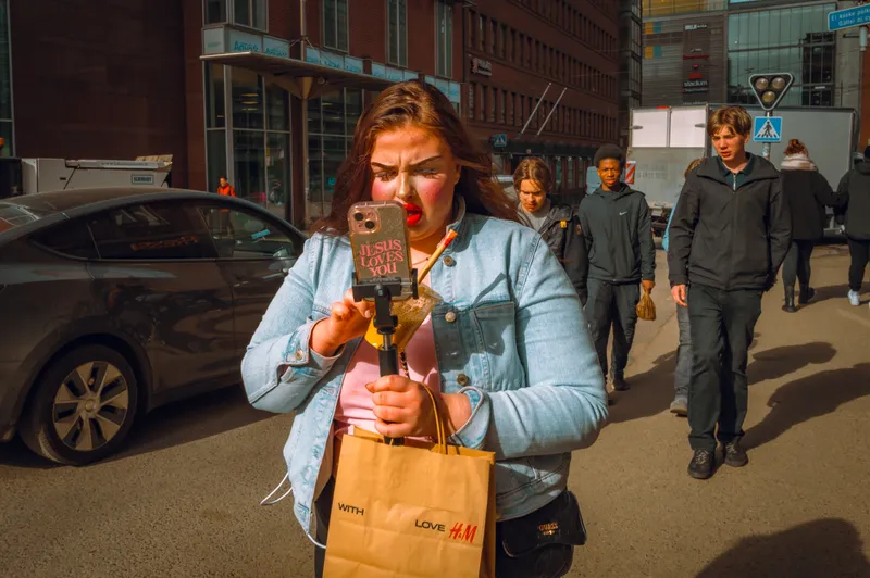 A young woman with bold red lipstick films herself on a selfie stick while walking a sunlit city street, teens trailing behind her.