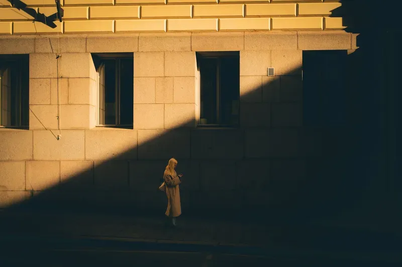 A woman checks her phone in a wedge of golden light against a stone building, a sharp shadow diagonal splitting the frame.