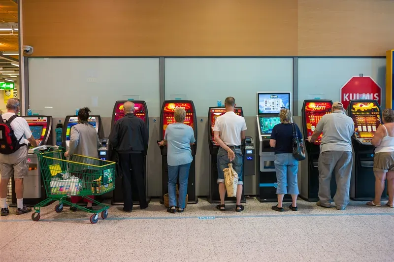 A row of people play slot machines lined against a wall in a supermarket lobby, a shopping cart parked among them.