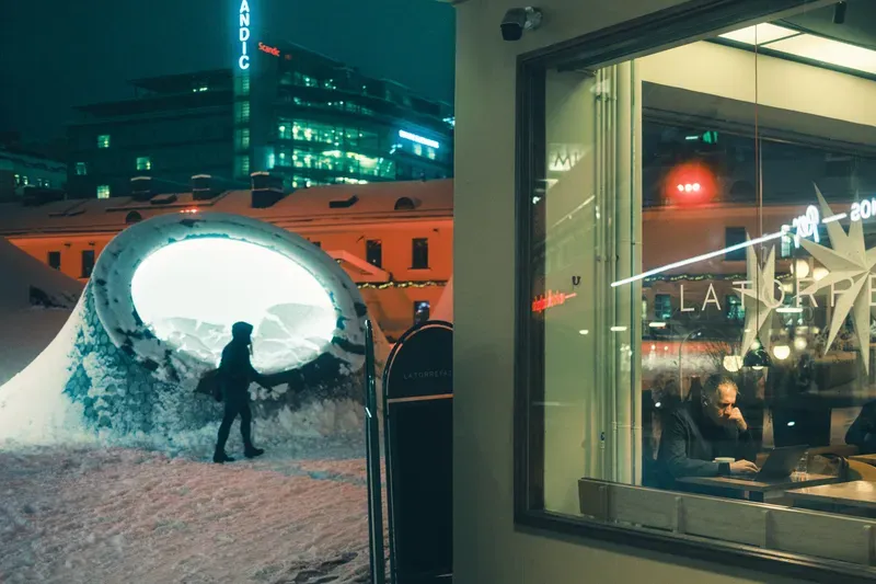 A figure walks through a glowing, snow-covered tunnel sculpture at night, a lit café window and a man working on his laptop visible beside it, city towers glowing in the distance.