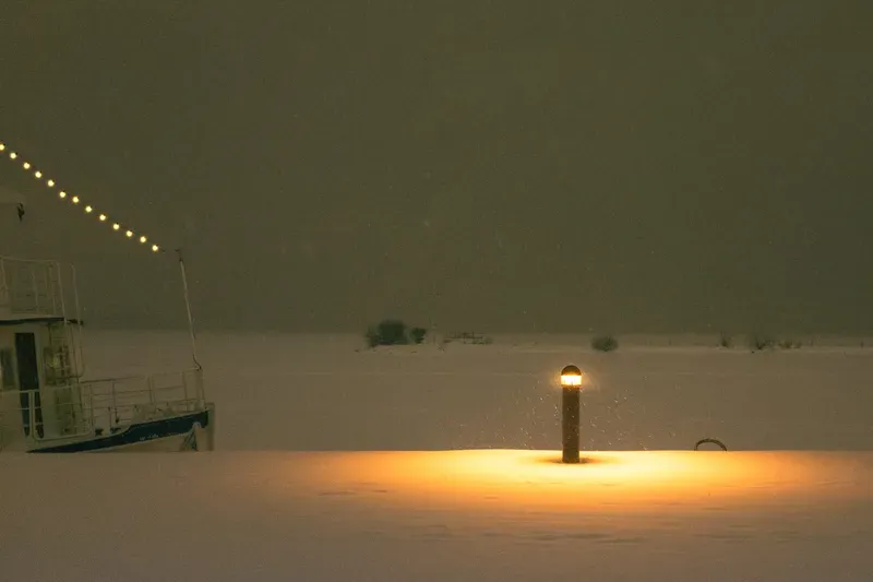 Snow falls at a winter marina where a glowing bollard lights the pier beside a moored boat strung with lights.