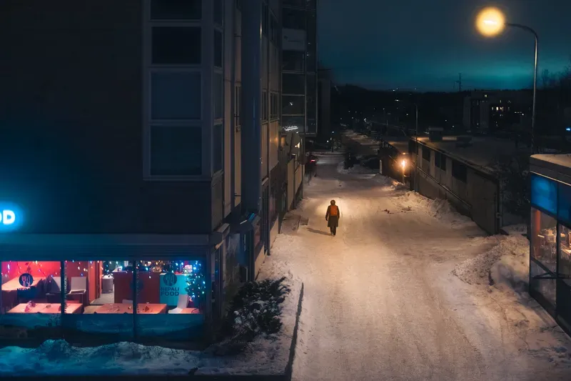A person in an orange coat walks alone down a snow-banked street at night, lit by a streetlamp beside a glowing Nepali restaurant sign.