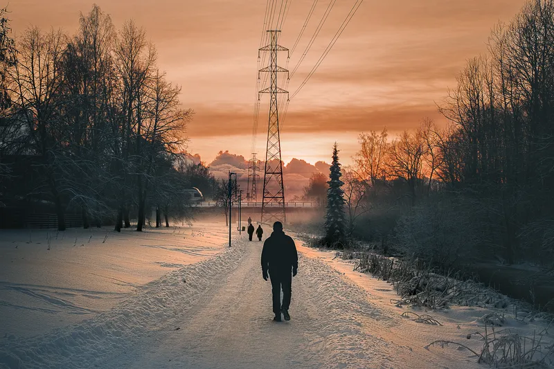 A man walks a snow-packed path toward a towering power line pylon, the low winter sun setting the sky ablaze in orange.