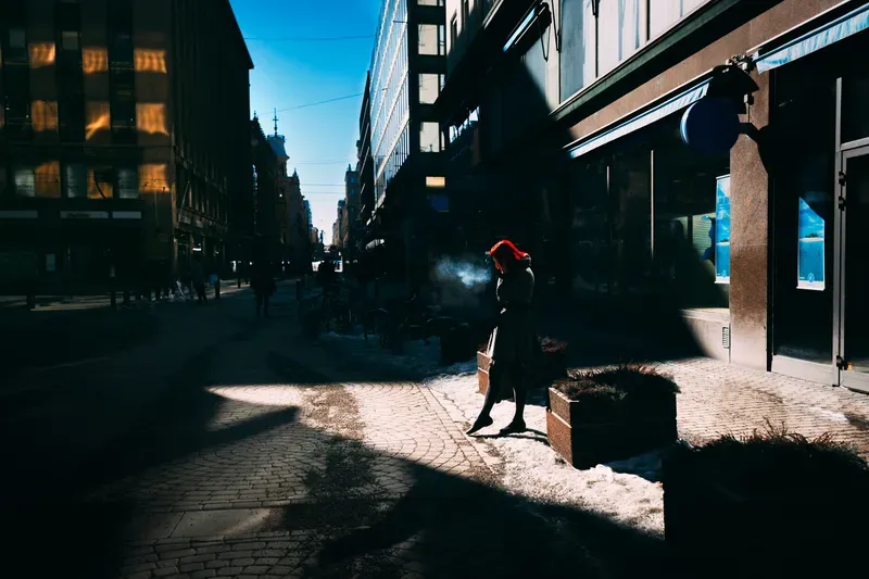 A figure in a red coat walks a snowy city street lined with long morning shadows.