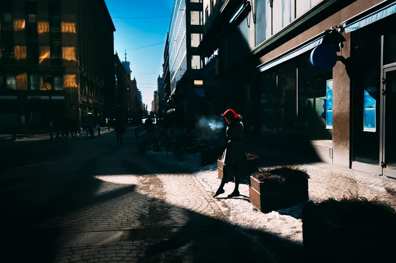 A figure in a red coat walks a snowy city street lined with long morning shadows.