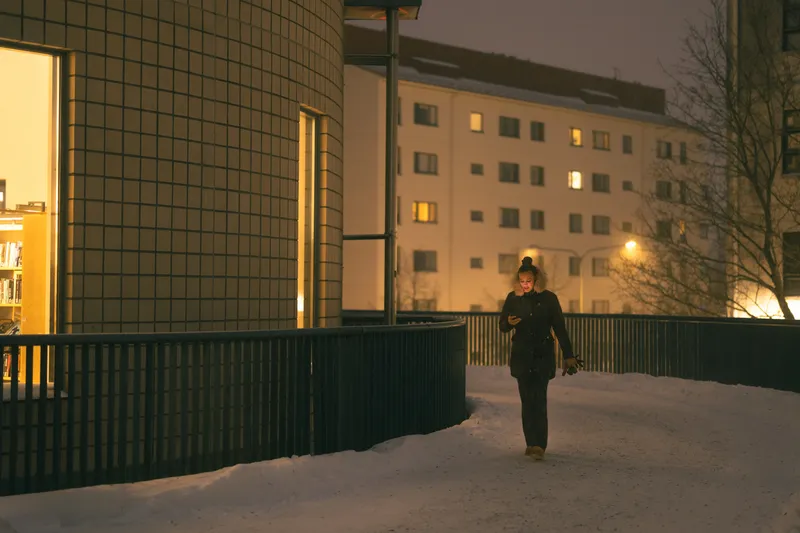 A woman checks her phone while walking along a snow-covered elevated walkway at dusk, apartment windows glowing behind her.