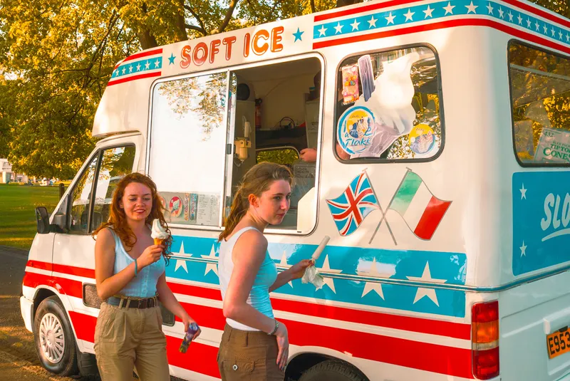 Two young women eat ice cream cones beside a red, white, and blue Soft Ice van under golden late-afternoon light.