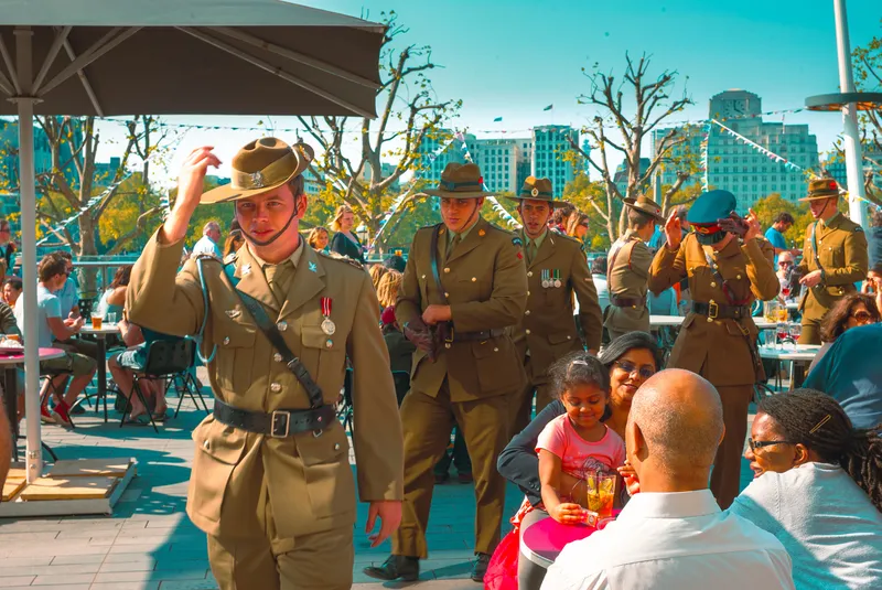 Soldiers in slouch hats and medals walk through a crowded riverside terrace lined with tables and bunting.
