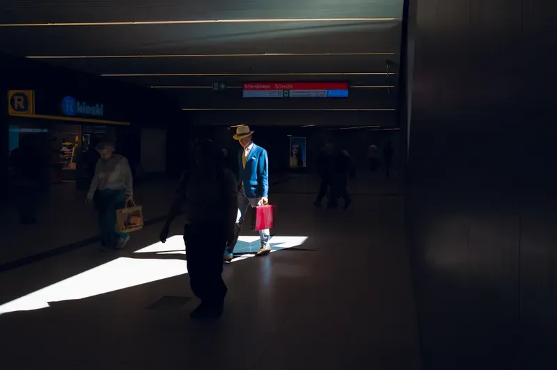 A man in a blue jacket and straw hat carries a red bag through a shaft of light in a metro station concourse, other travellers passing in the shadows.