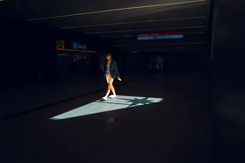 A man in a blazer and shorts crosses a near-black metro concourse through a single shaft of daylight, a compass star inlaid in the lit floor at his feet and a kiosk glowing faintly behind him.