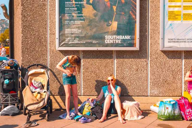 A girl and a woman in sunglasses sit against a wall beneath a Southbank Centre concert poster, surrounded by bags and beach toys.