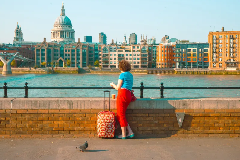 A woman in red trousers leans on a riverside wall beside a red suitcase, St Paul's Cathedral rising across the Thames.