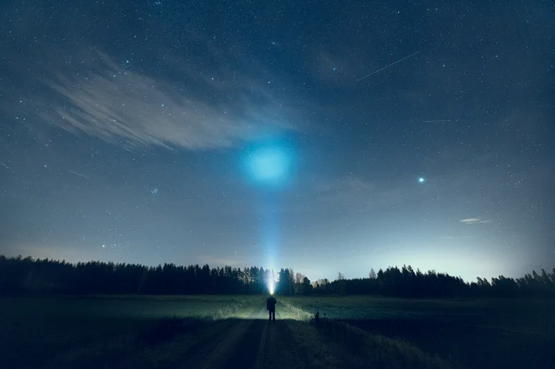 A lone figure stands on a dirt road beneath a glowing, star-filled night sky bordered by dark pine trees.