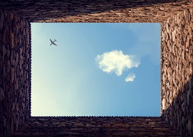 Looking straight up through a rough stone window frame at a blue sky with a passing airplane and a single cloud.