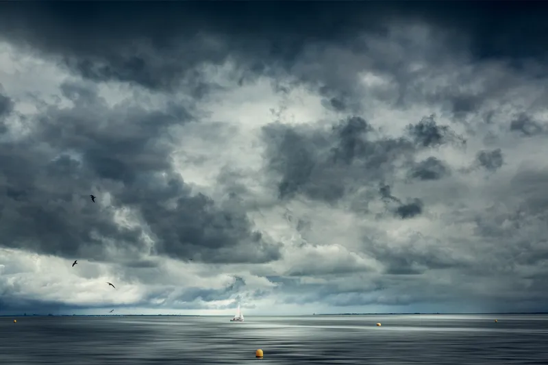 A single white sailboat drifts on calm water beneath a dramatic mass of dark storm clouds, birds circling below.