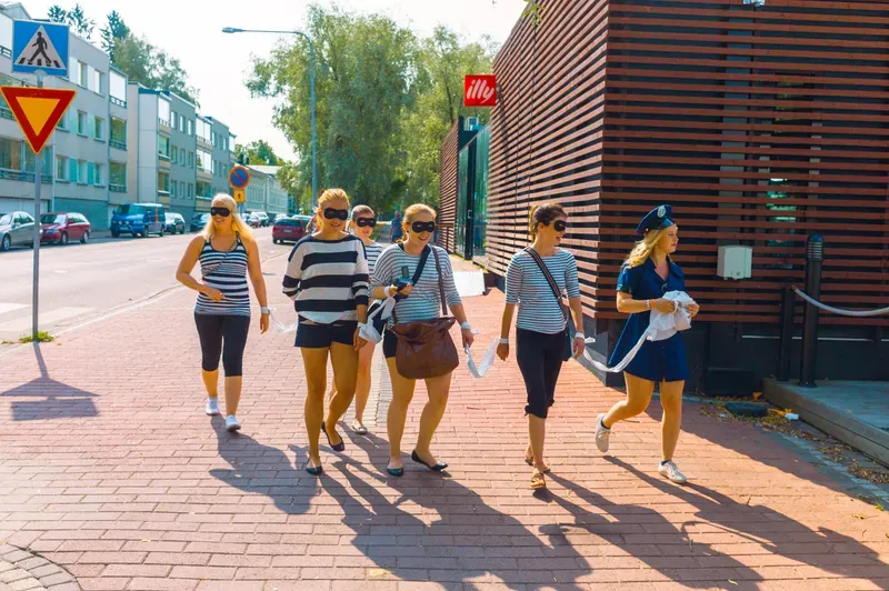 Four women in striped tops and black eye masks walk along a sunlit brick pavement, a fifth in a police-style cap and navy dress hurrying beside them with a white cloth.
