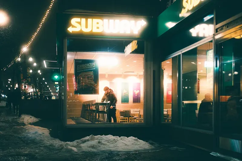 A man stands inside a brightly lit Subway sandwich shop at night, seen through the storefront window from the snowy street outside.