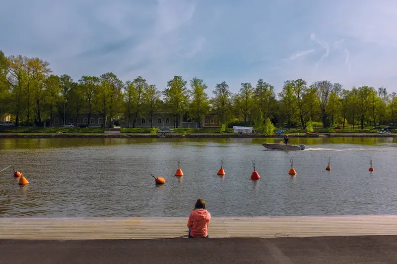 A woman in a coral jacket sits alone at the edge of a wooden deck facing a canal, a line of orange mooring buoys on the water and a small boat passing a row of summer trees.
