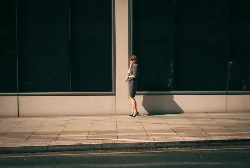 A woman in a gray dress leans against a sunlit pillar between dark glass storefronts, checking her watch.