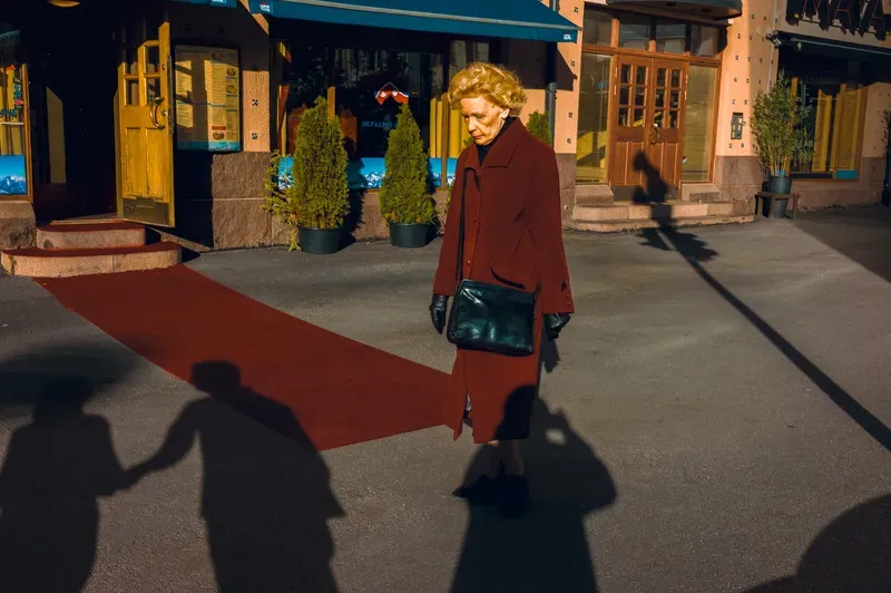 A woman in a burgundy coat and leather bag walks across a red carpet outside a restaurant, long shadows of onlookers stretching across the pavement.