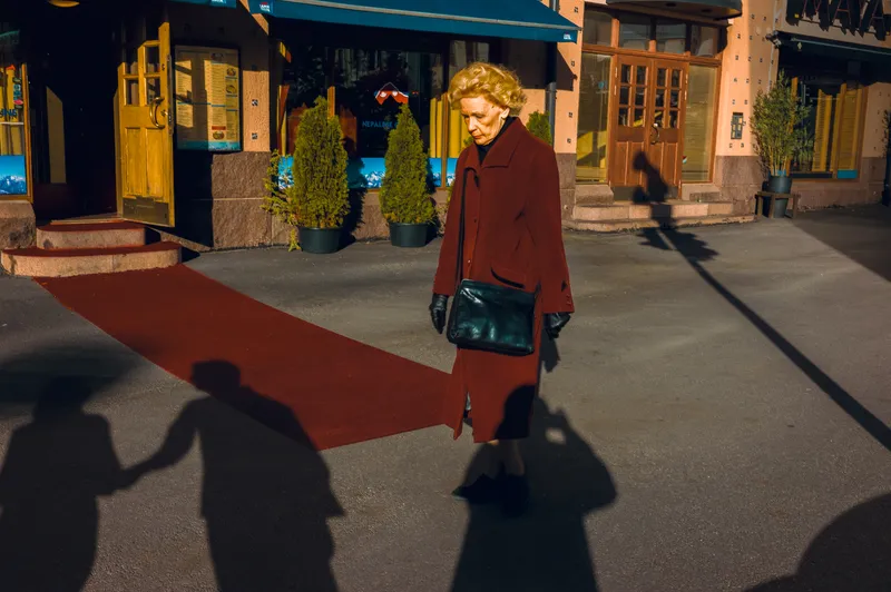 A woman in a burgundy coat and leather bag walks across a red carpet outside a restaurant, long shadows of onlookers stretching across the pavement.