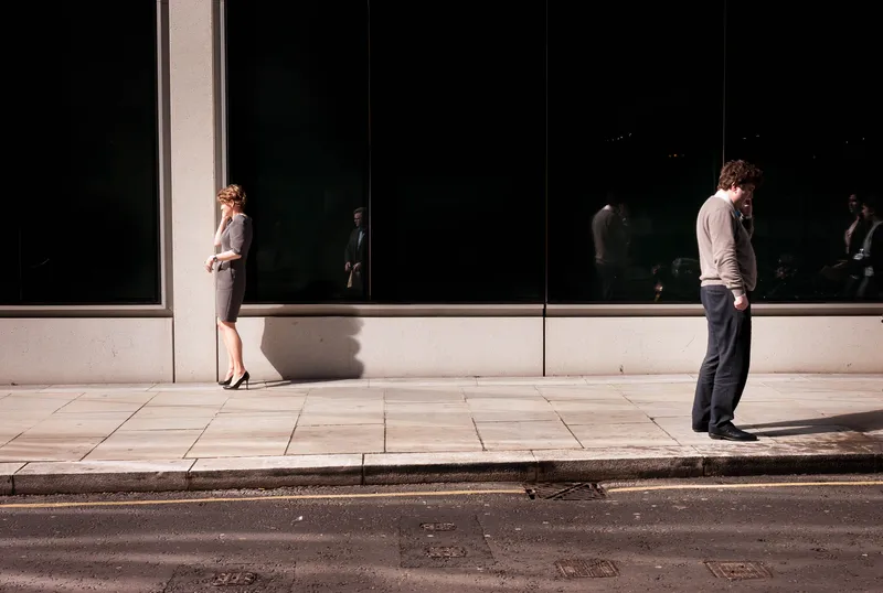 A woman in a gray dress leans against a sunlit pillar while a man on his phone stands apart on the opposite curb, dark glass storefronts behind them.