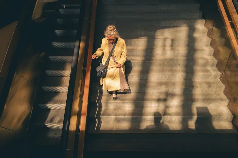 Overhead view of an elderly woman in a cream coat descending sunlit stairs beside an escalator, long shadows stretching across the steps.