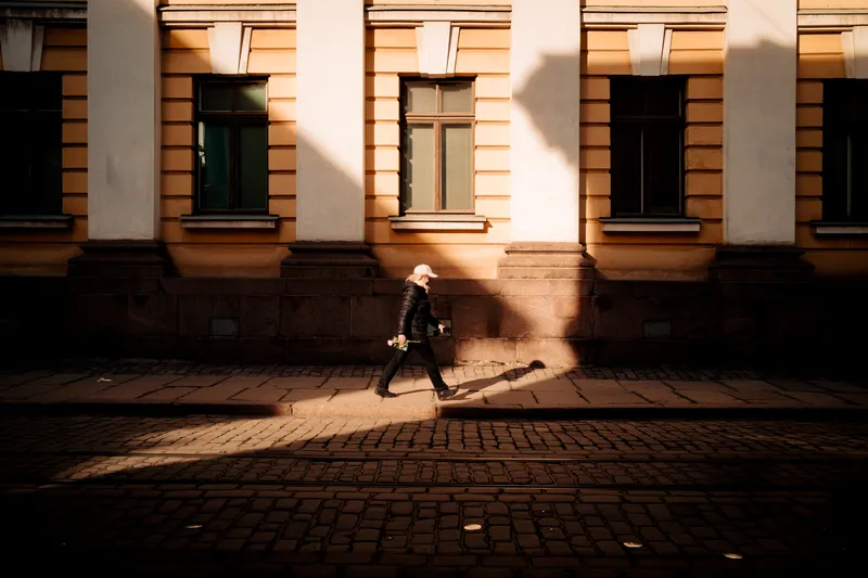 A woman in a cap walks along a sunlit cobblestone street holding a flower, her long shadow trailing across an ornate yellow building facade.