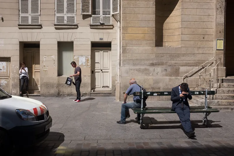 People pause on a sunlit street of shuttered stone buildings, two men seated on a bench and a woman leaning in a doorway beside a parked car.