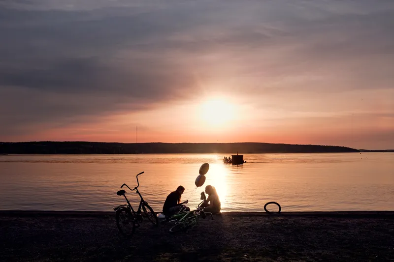 Two kids with bicycles and balloons sit at the water's edge, silhouetted against a glowing pink and orange sunset over the lake.