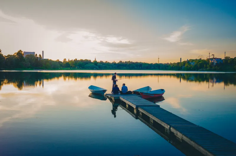 Friends gather at the end of a wooden dock with rowboats moored alongside, fishing as the sky glows gold over the city skyline.