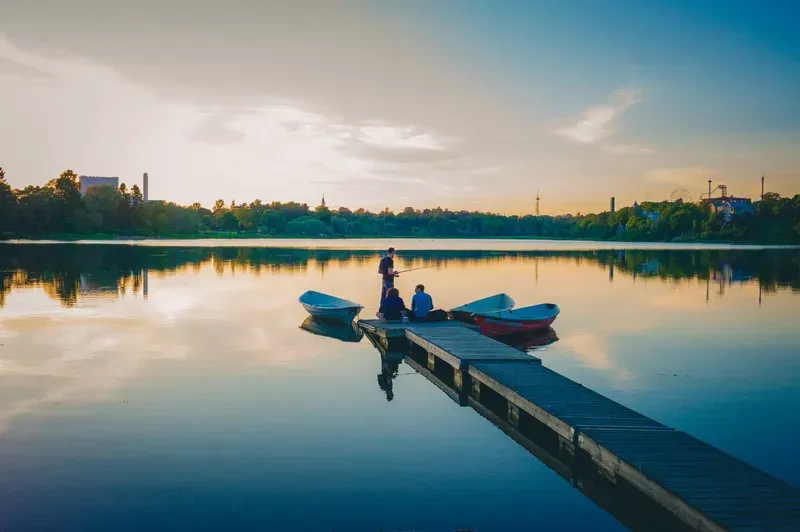 Friends gather at the end of a wooden dock with rowboats moored alongside, fishing as the sky glows gold over the city skyline.