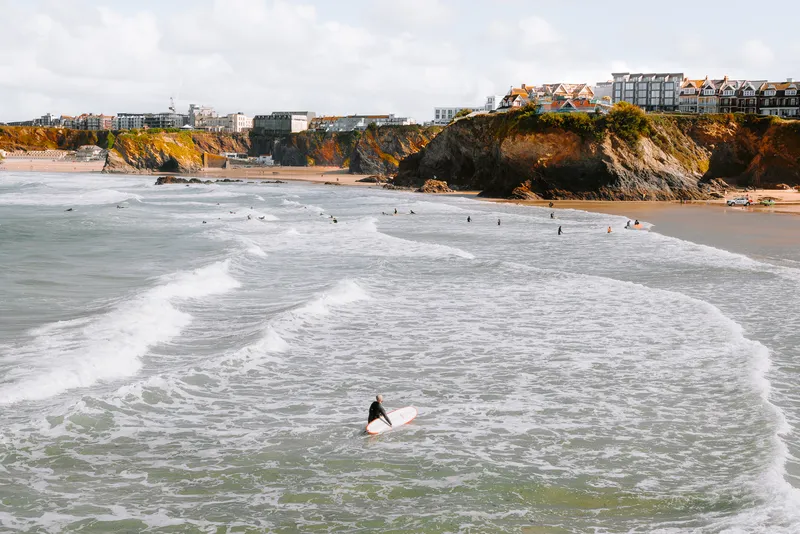 A surfer wades into the waves carrying their board, cliffs and a seaside town rising behind them under an overcast sky.
