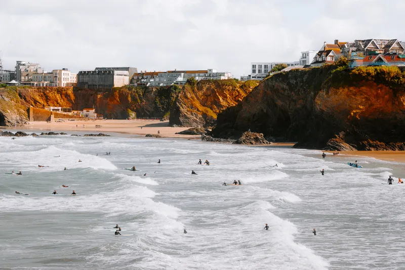 Dozens of surfers wait and ride waves off a sandy beach, cliffside houses and a seaside town spread out above the shore.