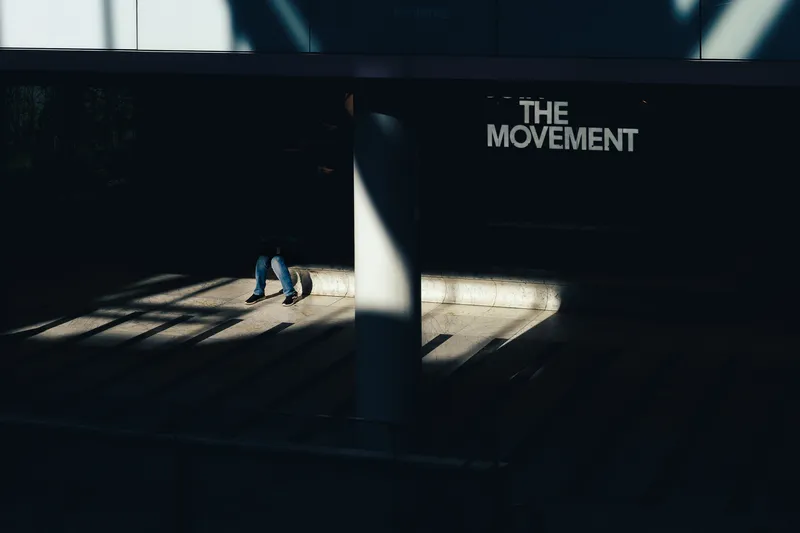 A seated figure's legs are lit by a stray beam of light in an otherwise dark hall beneath a sign reading 'THE MOVEMENT'.